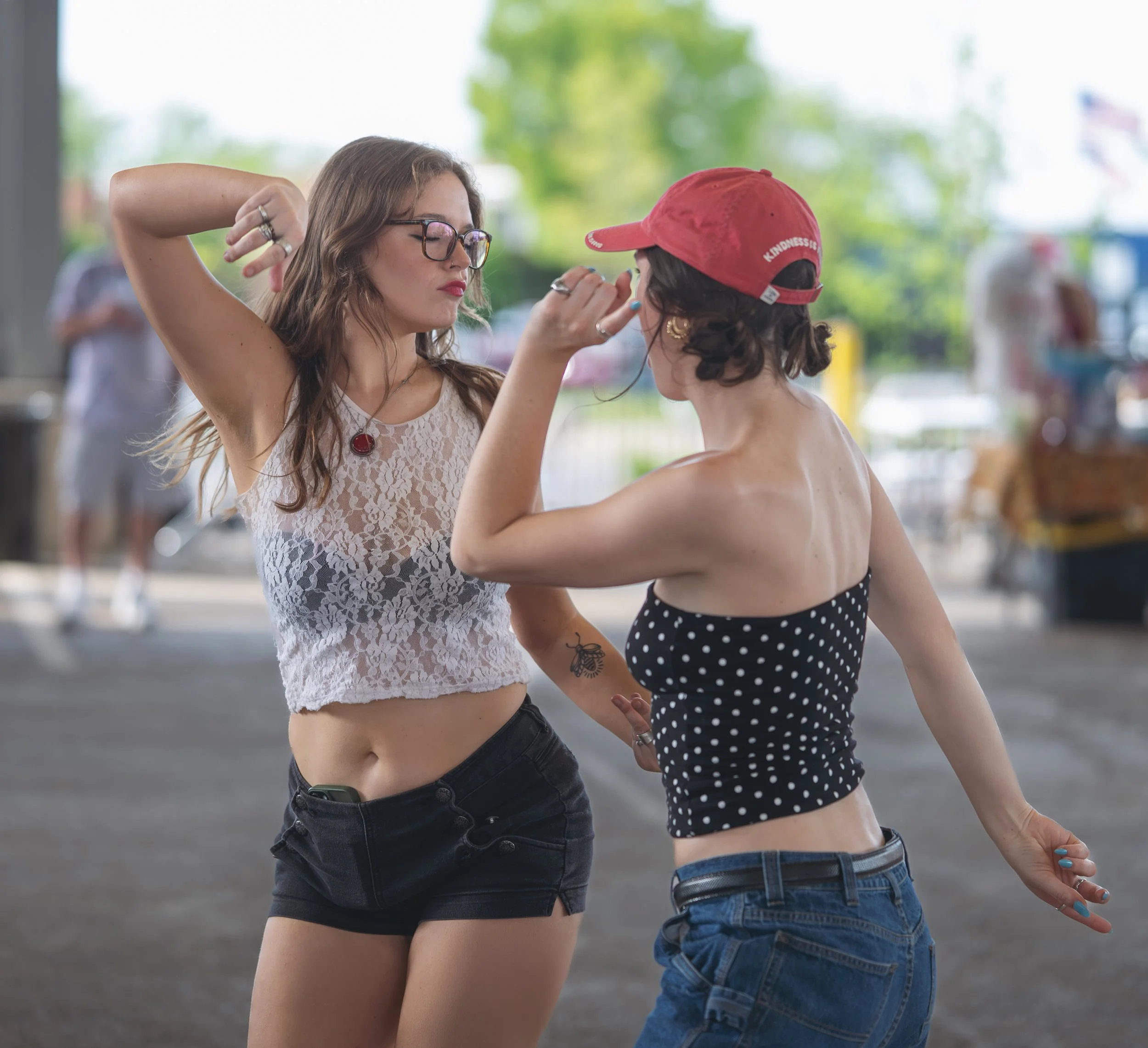 Two women are dancing outdoors, facing each other. The woman on the left has long curly hair, glasses, and is wearing a white lace top and black shorts. The woman on the right has curly hair, a red cap, and is dressed in a black polka dot crop top an