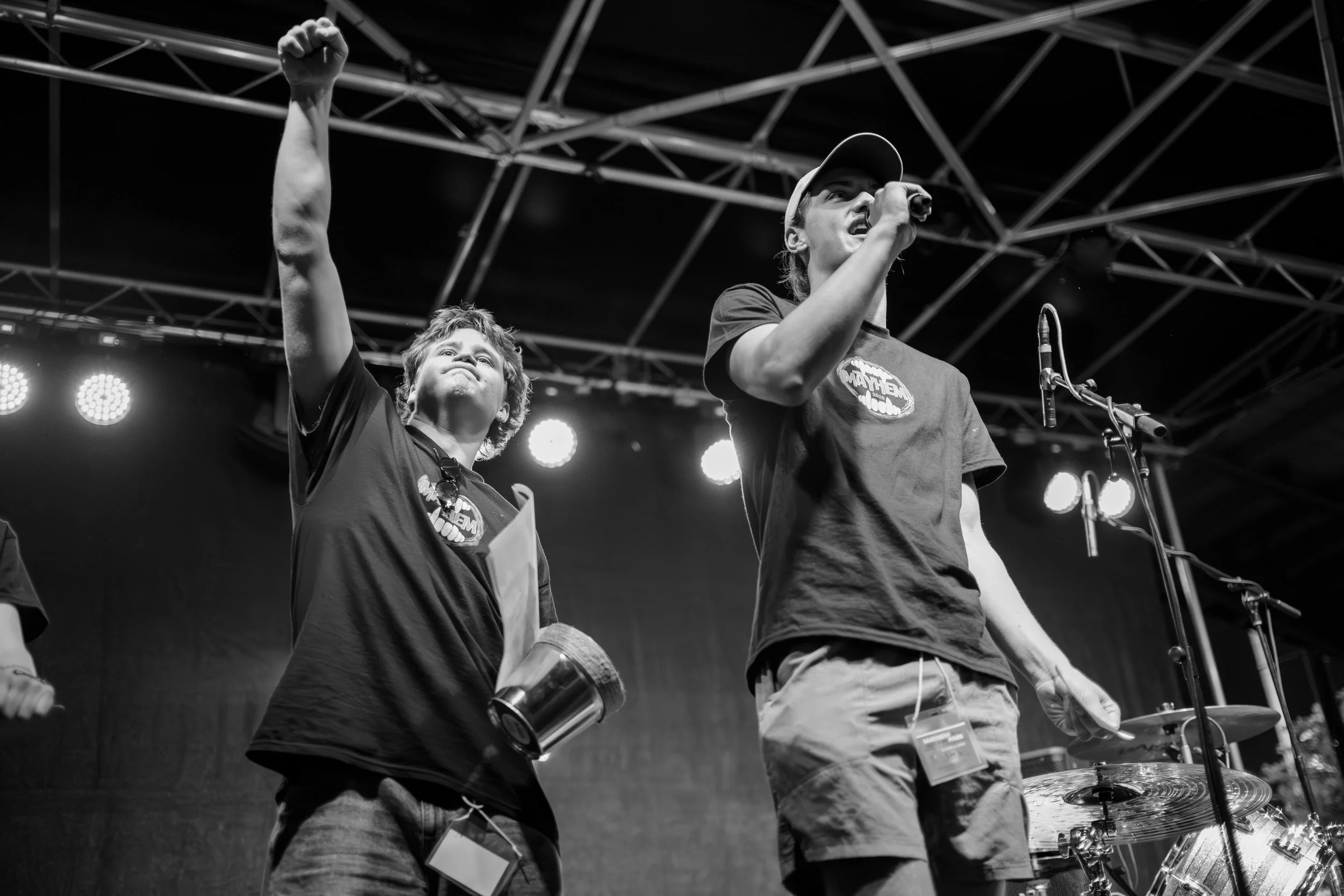 Two young men on stage at the Mayhem music festival, one speaking into a microphone and the other raising fist, both wearing t-shirts with a logo, with stage lights overhead.