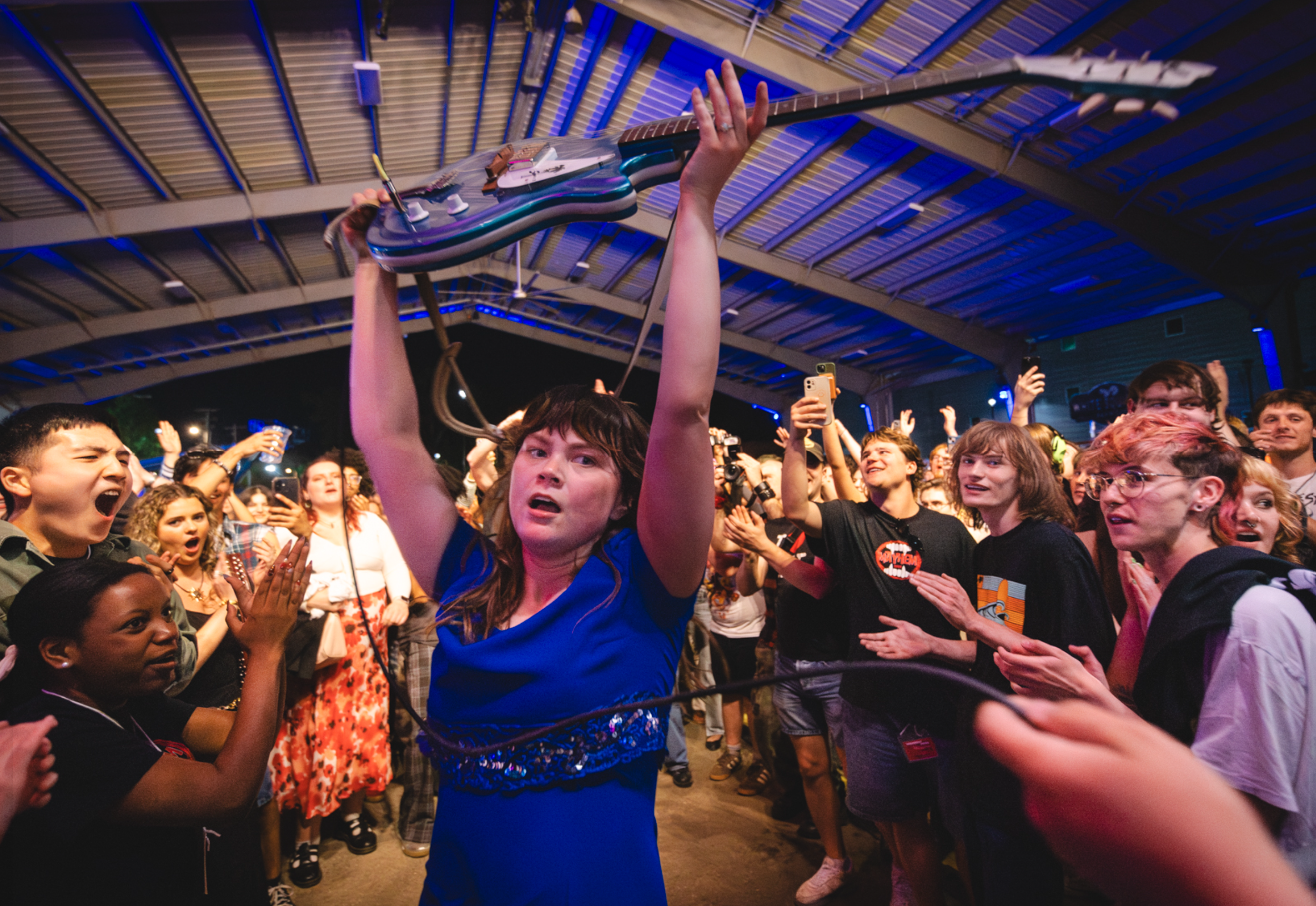 A crowd of excited people at a concert or event, with a woman holding an electric guitar above her head in the center, under a large, curved roof structure with blue lighting.