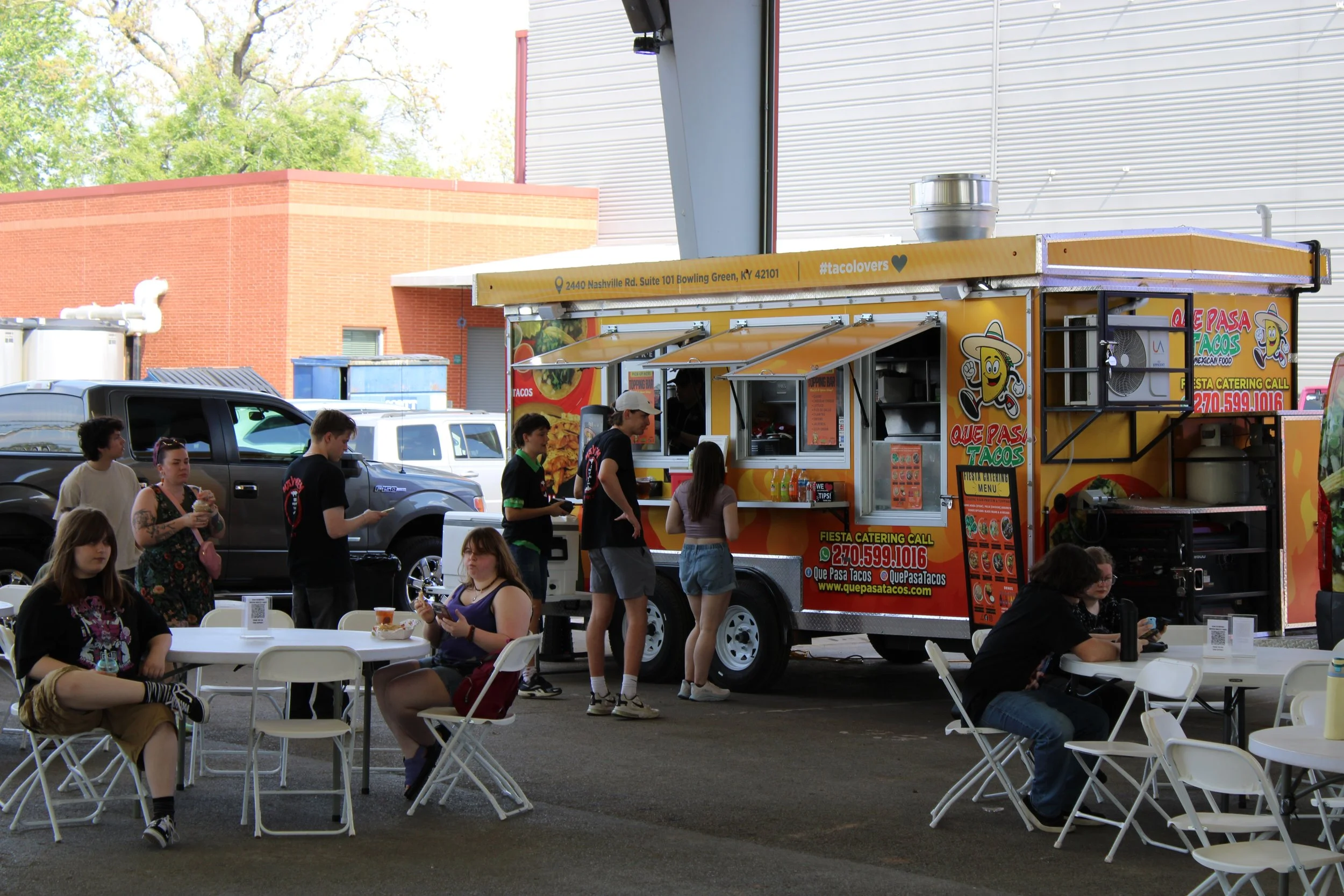 People lining up and sitting at tables outside at a Que Pasa Mexican food truck, with a brick building and trees in the background.