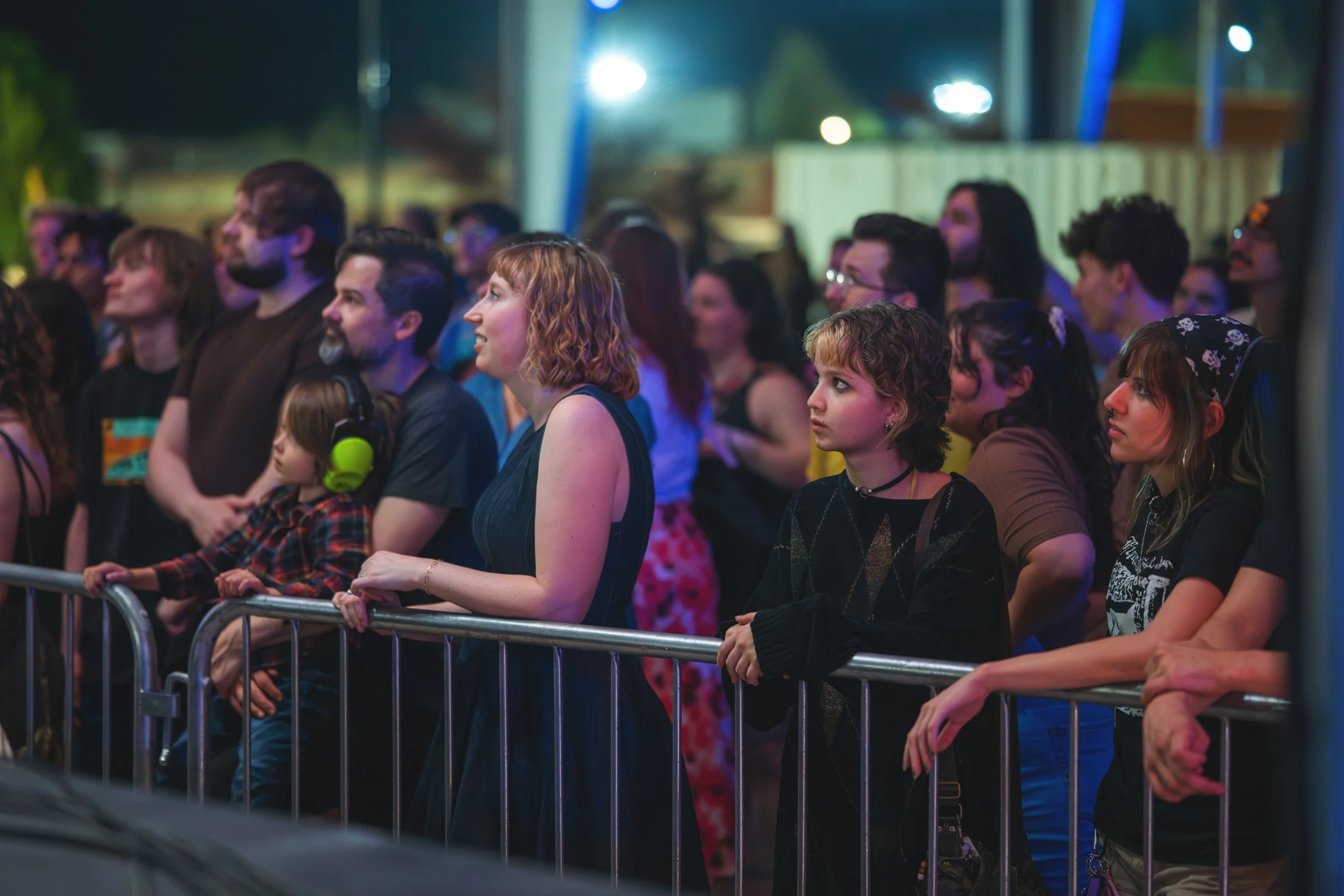 A diverse group of people attending the Mayhem music festival relaunch, standing behind a metal barrier, listening and watching attentively in a night setting with blue and purple lighting.