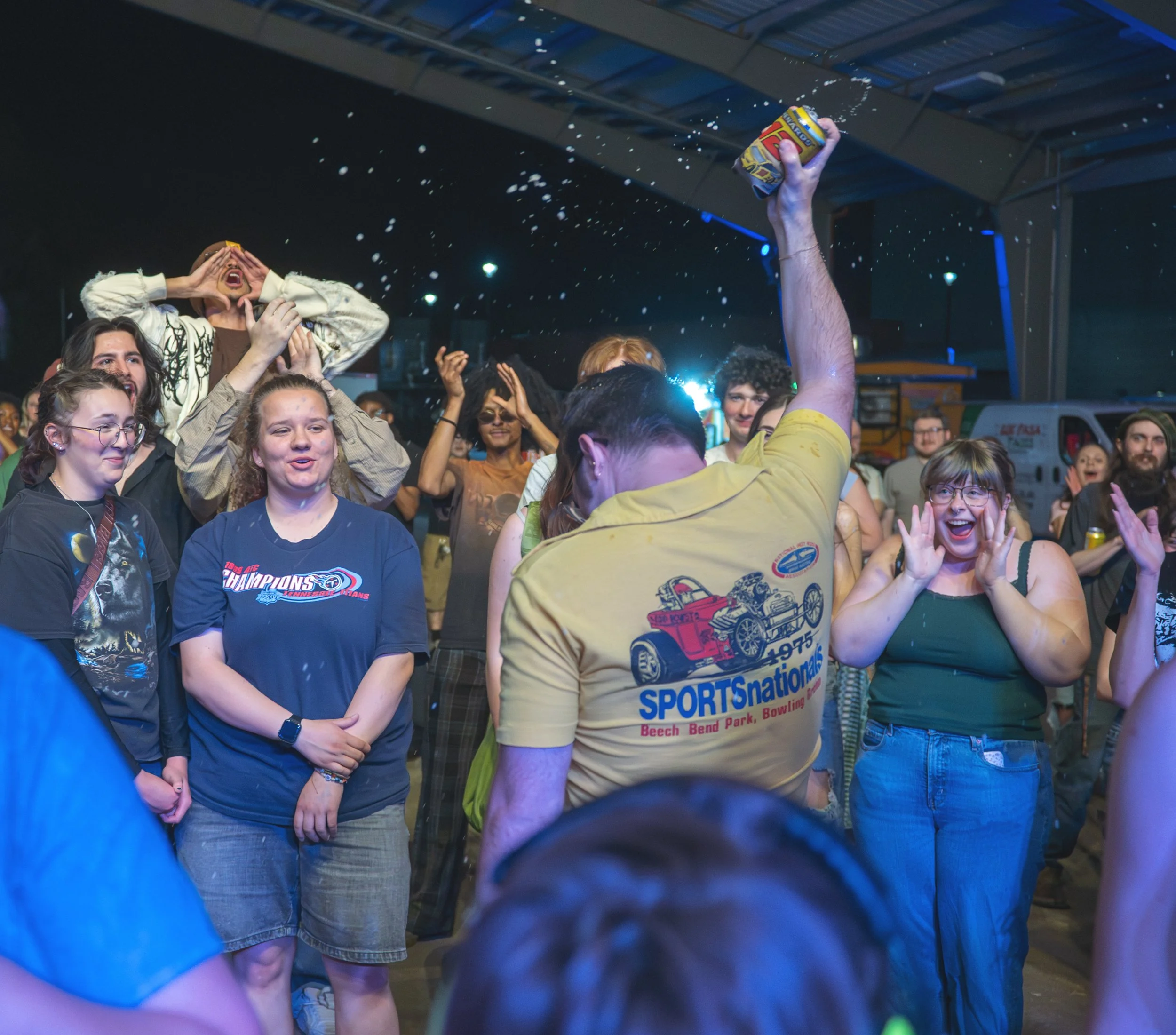 A group of people gathered outdoors at night, some are smiling and looking towards the camera. One person is holding a can, and another is shouting or cheering with hands around their mouth. The crowd appears to be enjoying Mayhem 2026.