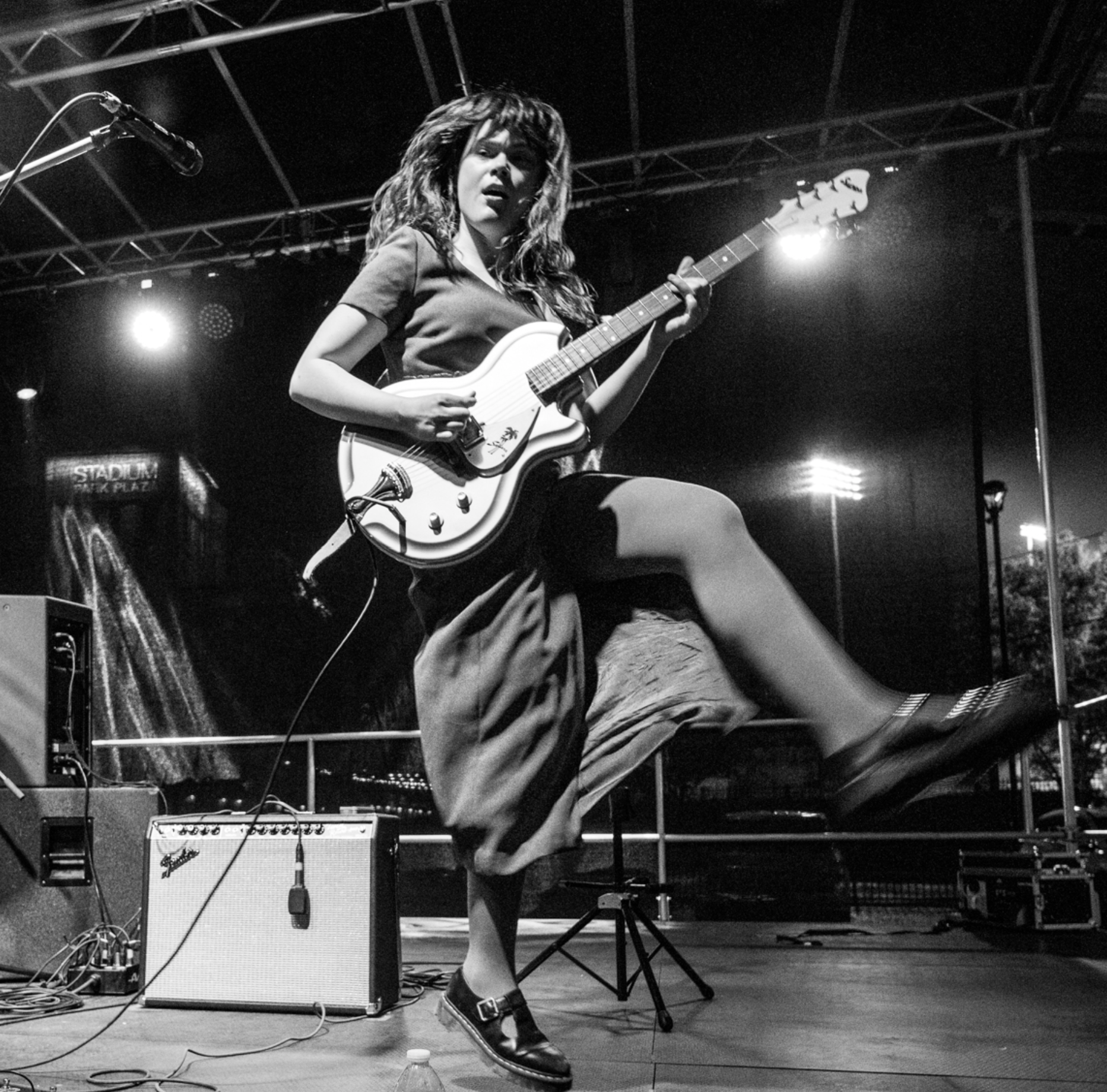 A woman with curly hair playing an electric guitar on stage, with stage lights and sound equipment visible.