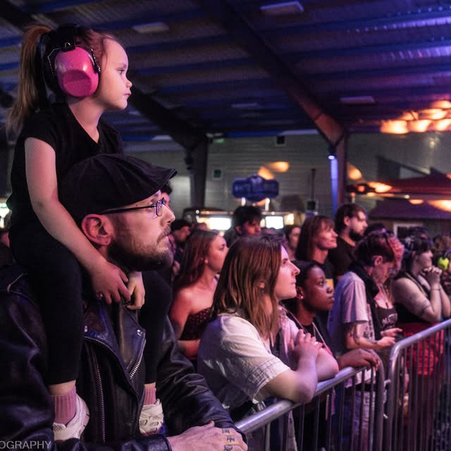A young girl wearing pink headphones sits on a man's shoulders at a concert or event, both attentively watching the stage, surrounded by a crowd of diverse spectators in an indoor venue.