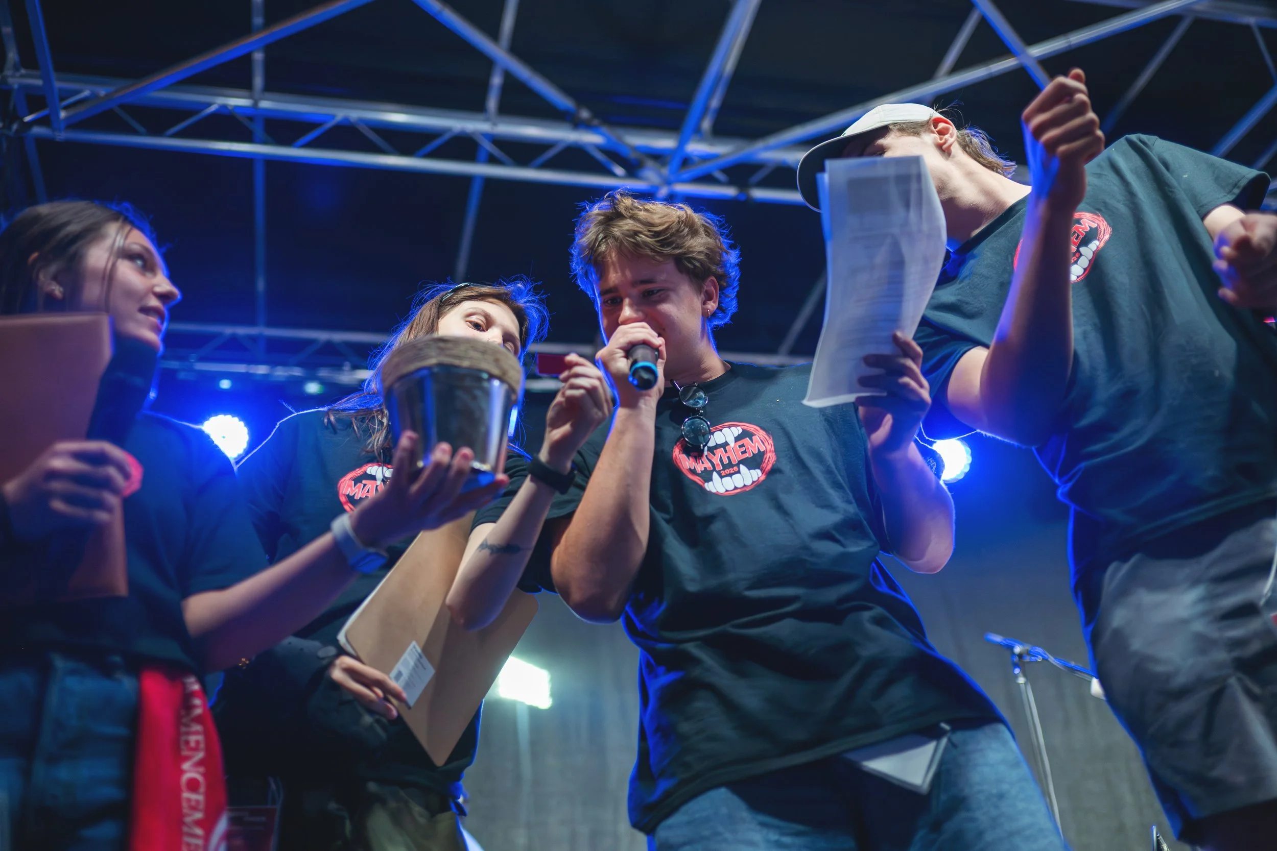 Group of young people on stage at a concert or event with one person holding a microphone and reading from a paper, wearing black T-shirts with a red and white 'Mayhem' logo.
