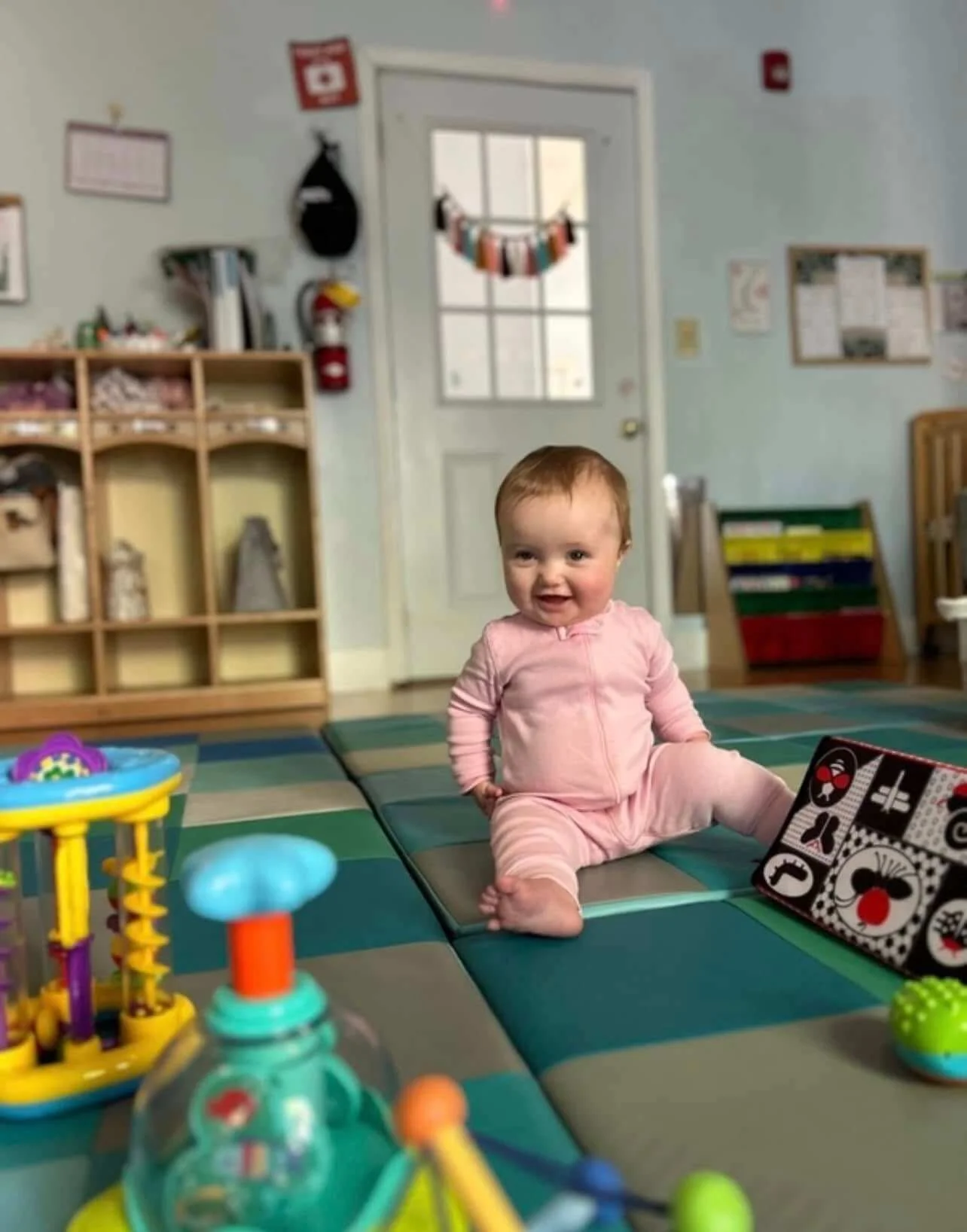 A toddler sitting on colorful mats in a playroom, smiling and wearing a pink zip-up outfit, surrounded by toys.