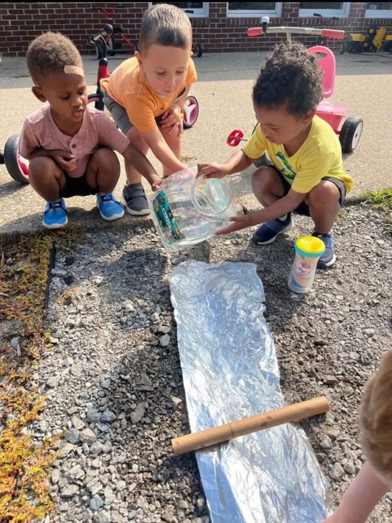 Three children squatting outside on gravel, pouring water from a plastic jug onto a reflective foil sheet, with tricycles and a brick building in the background.