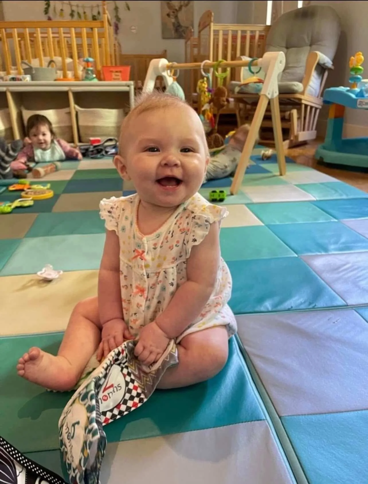 Smiling baby girl sitting on colorful foam mats in a playroom, with other children and toys in the background.