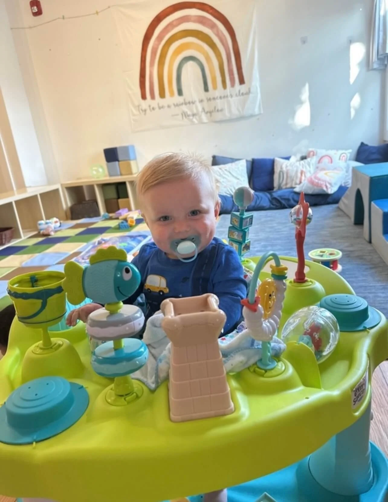 Young child with blonde hair and pacifier in mouth sitting in a colorful activity center, smiling at the camera. The background features blue and white cushions, a rainbow poster on the wall, and a cozy indoor environment.