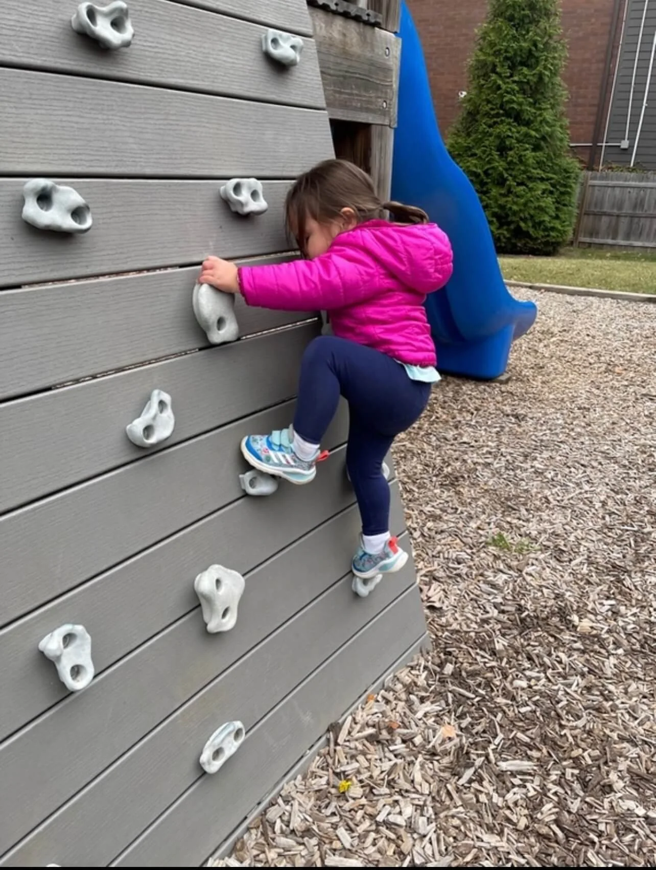 A young girl in a pink jacket, navy pants, and sneakers climbing an outdoor play climbing wall.