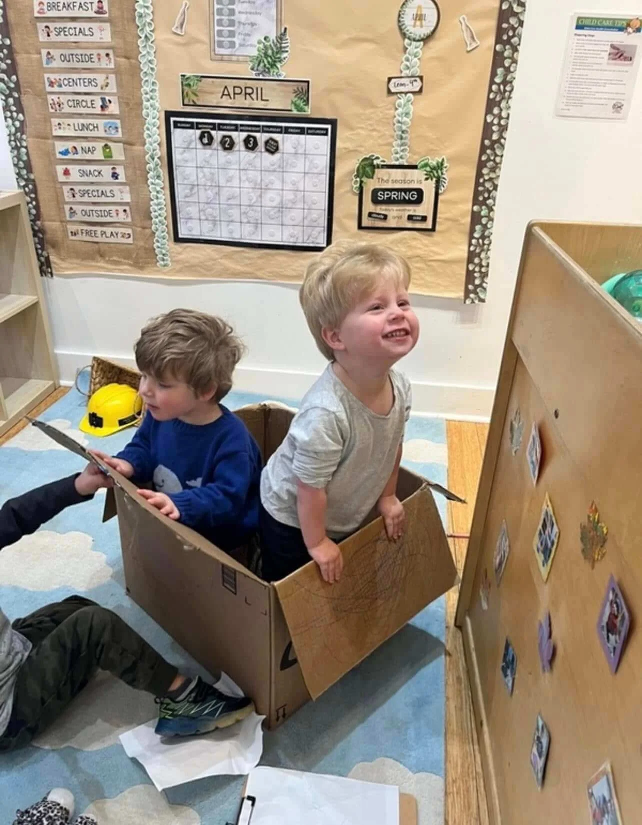 Two young boys are sitting in a cardboard box in a classroom, with one boy smiling and looking up, and the other boy looking down at a paper or book. The classroom has a bulletin board with calendar and schedule items, and a yellow construction helme