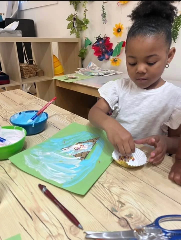 A young girl working on a craft project at a wooden table, pasting colorful sprinkles onto a paper sailboat on a painted blue background.