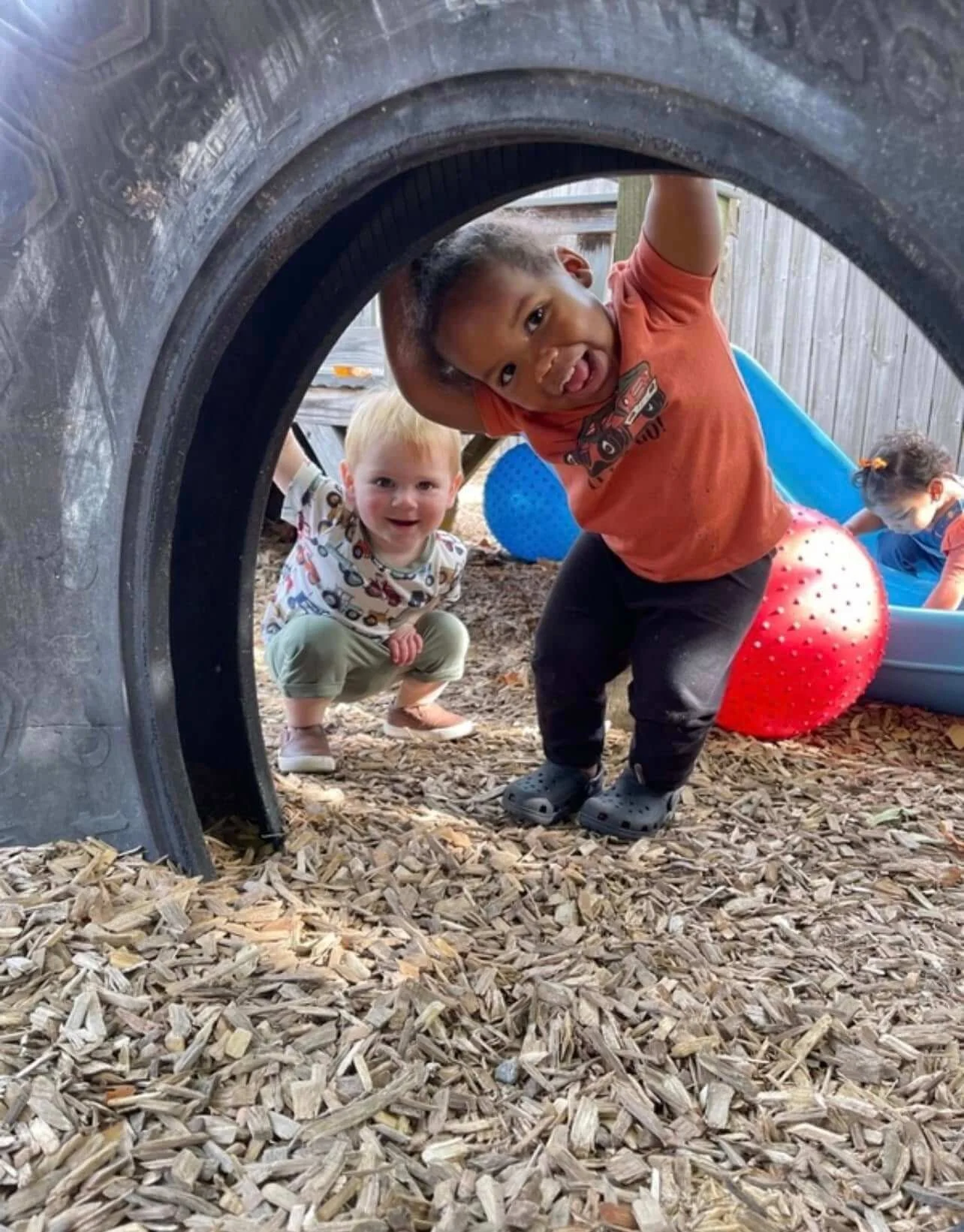 Two young children playing outside under a large tire, with one boy climbing through and another squatting nearby, both smiling and enjoying their time.