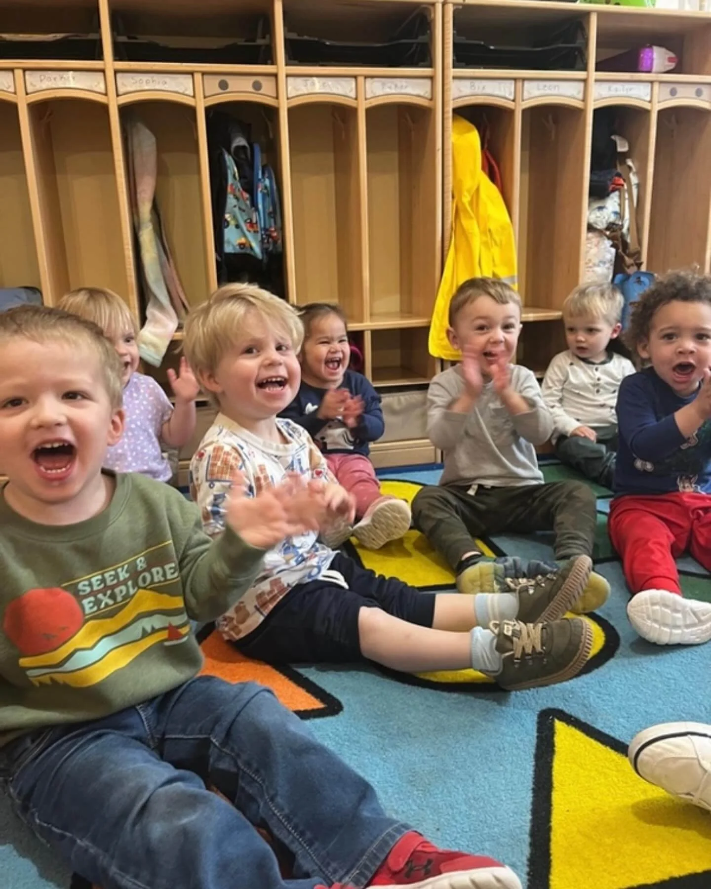 A group of young children sitting on a colorful carpet in a classroom, laughing and clapping.