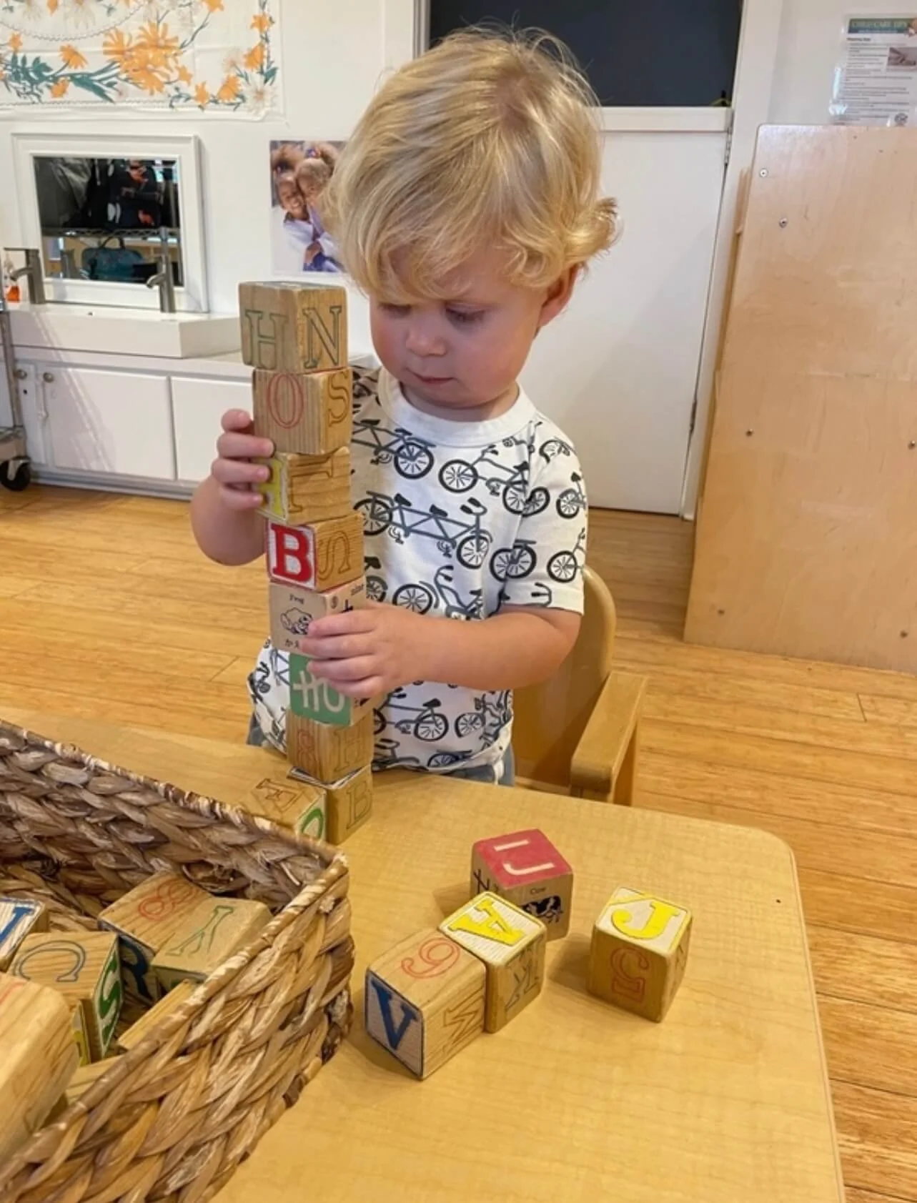 A young boy with blonde curly hair stacking colorful wooden blocks with letters on a wooden table in a room with wooden floors and white furniture.