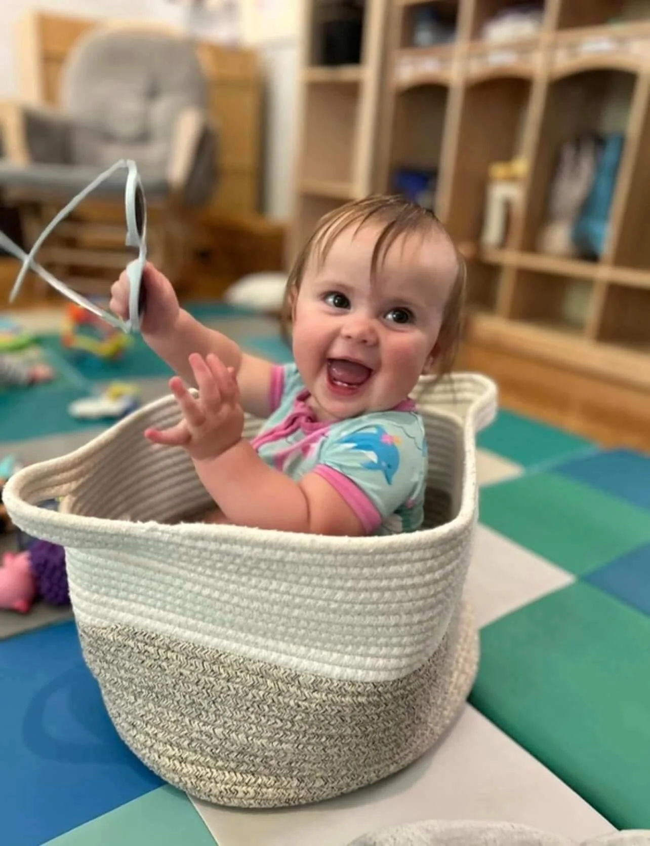 A happy baby sitting in a woven basket, holding a pair of glasses, in a room with wooden shelves and colorful foam mats on the floor.