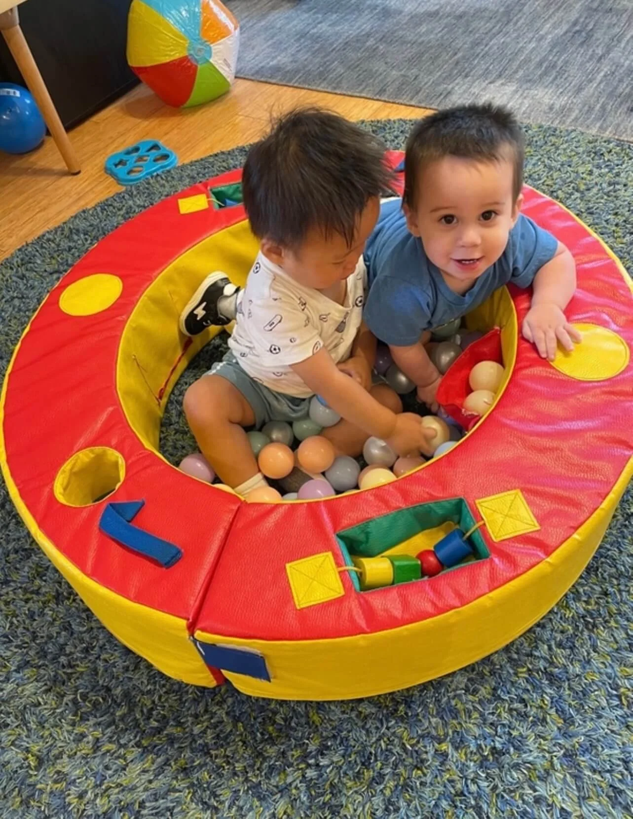 Two young children playing in a small ball pit, sitting on a multicolored padded border with a blue carpet underneath.