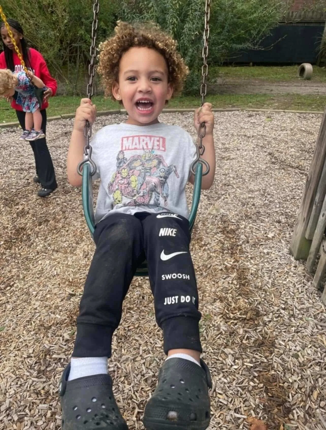 A young boy with curly hair on a swing, smiling widely at the camera, at a playground with wood chip ground, with a woman and a girl in the background.