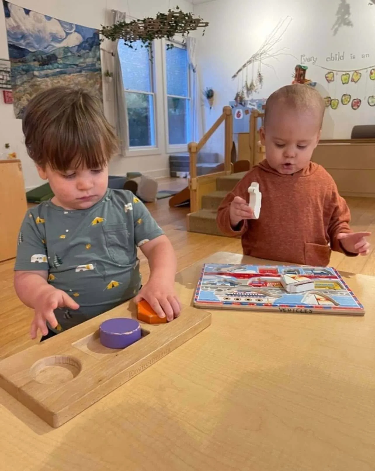 Two young children playing at a table with toys and a puzzle in a room with windows and wall decorations
