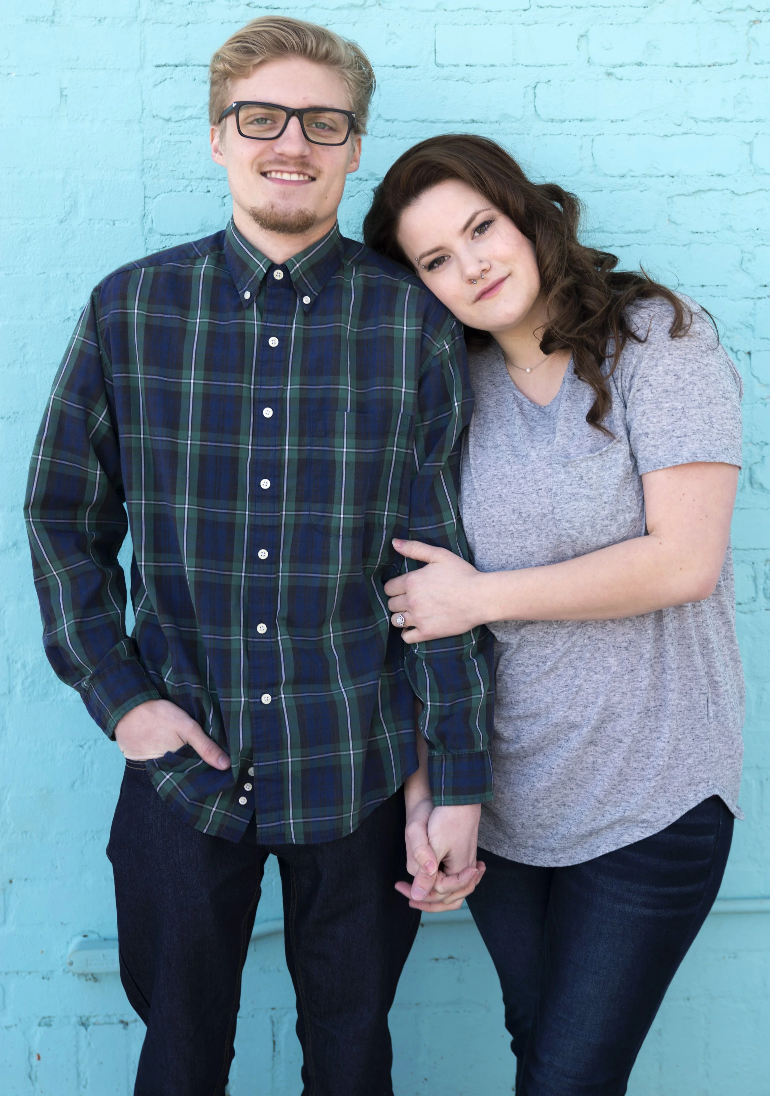 A young man and woman stand closely together in front of a blue brick wall, holding hands and smiling.