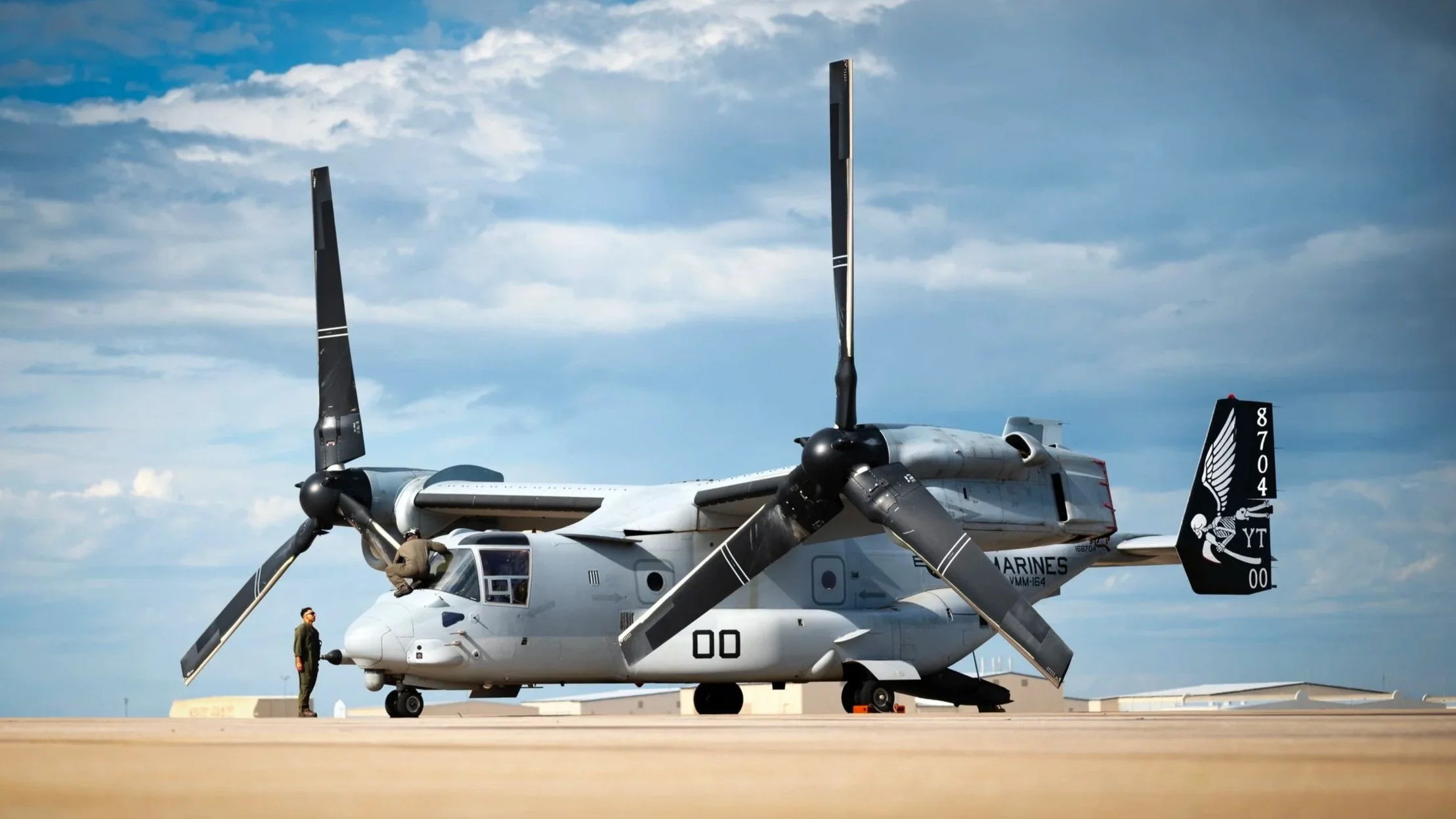 A military tiltrotor aircraft known as an Osprey with its rotors in horizontal position parked on the ramp, with a person standing near the front and another person near the rotors, under a cloudy sky.