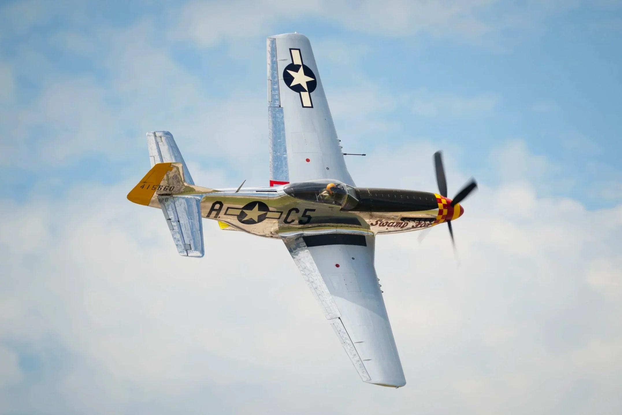 A P-51 Mustang airplane flying in cloudy sky with its nose painted red and yellow checkered pattern, and military insignia on the wings and fuselage.