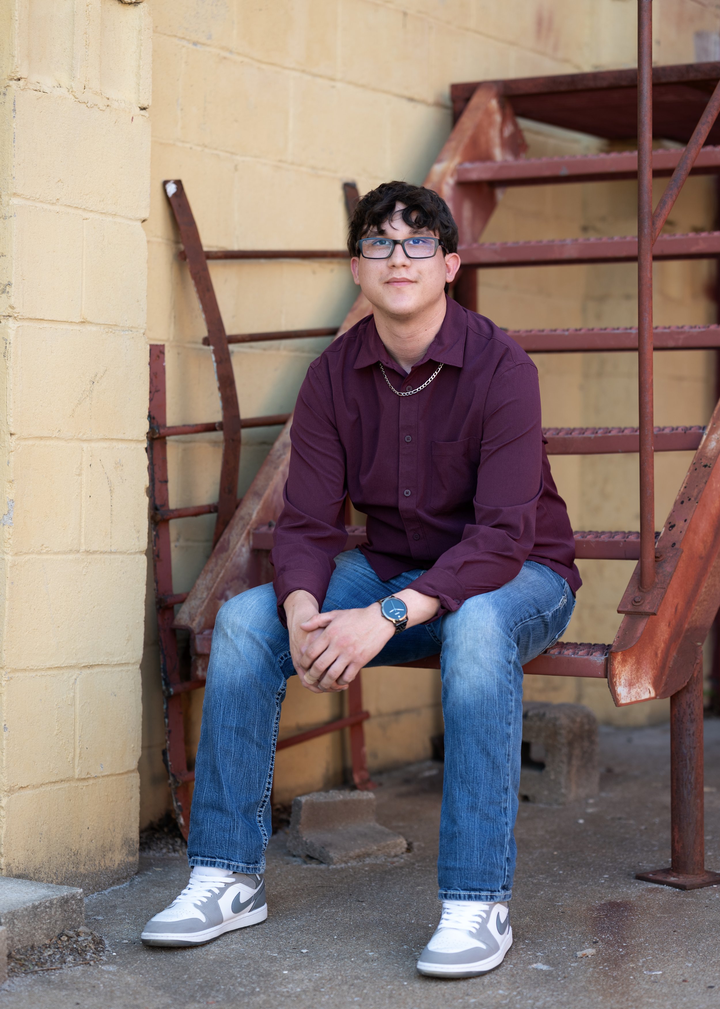 A young man with dark curly hair, glasses, and light skin, sitting on a metal staircase outdoors against a yellow brick wall, wearing a maroon shirt, blue jeans, white and gray Nike sneakers, a watch, and a silver chain necklace.