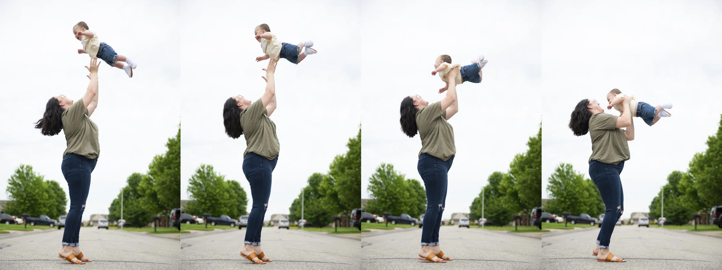 A woman with dark, shoulder-length hair wearing a green t-shirt, ripped jeans, and brown sandals jumping in the air, holding a young boy in a cream shirt and denim shorts upward. The scene is outdoors on a surrounding street with trees and parked car