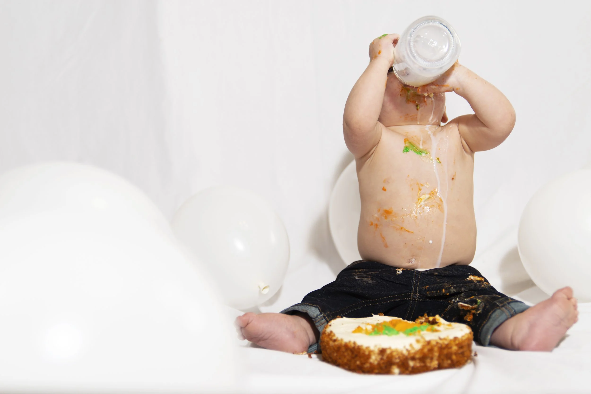 Baby sitting among white balloons with birthday cake and a partially eaten cake on the floor, drinking from a bottle while messy with cake and frosting.