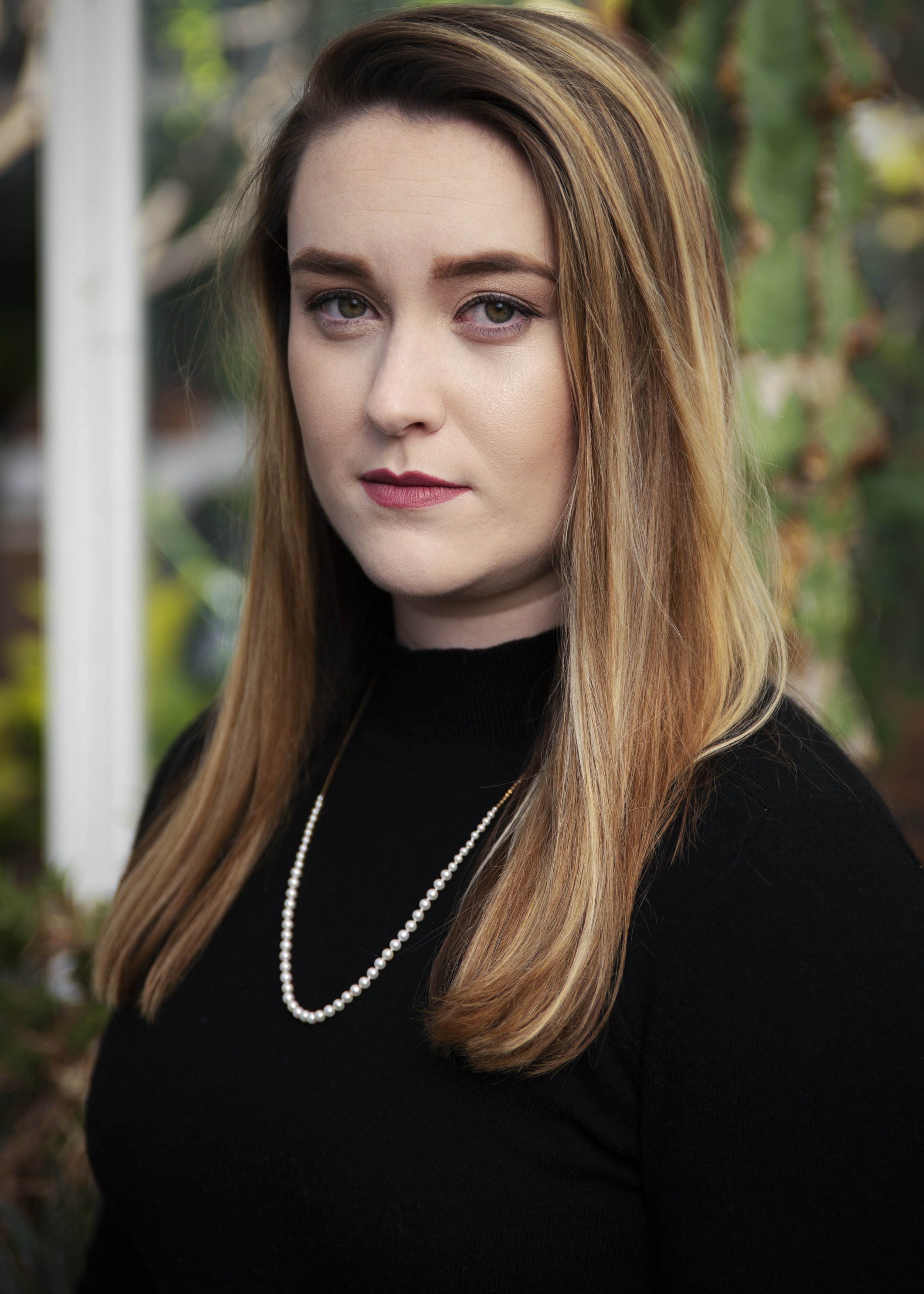 A woman with long light brown hair wearing a black top and pearl necklace, standing outdoors with greenery in the background.