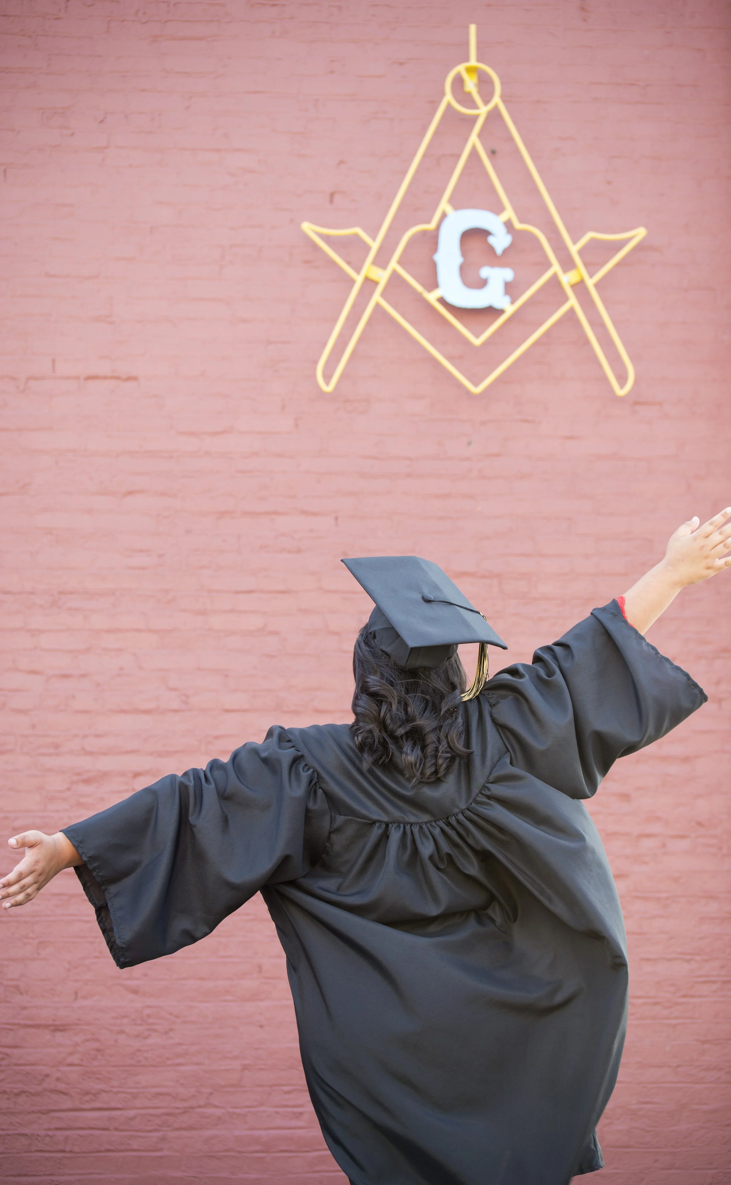 Person in graduation gown and cap dancing or celebrating in front of a pink brick wall with a neon Masonic square and compass symbol, and a white letter G.