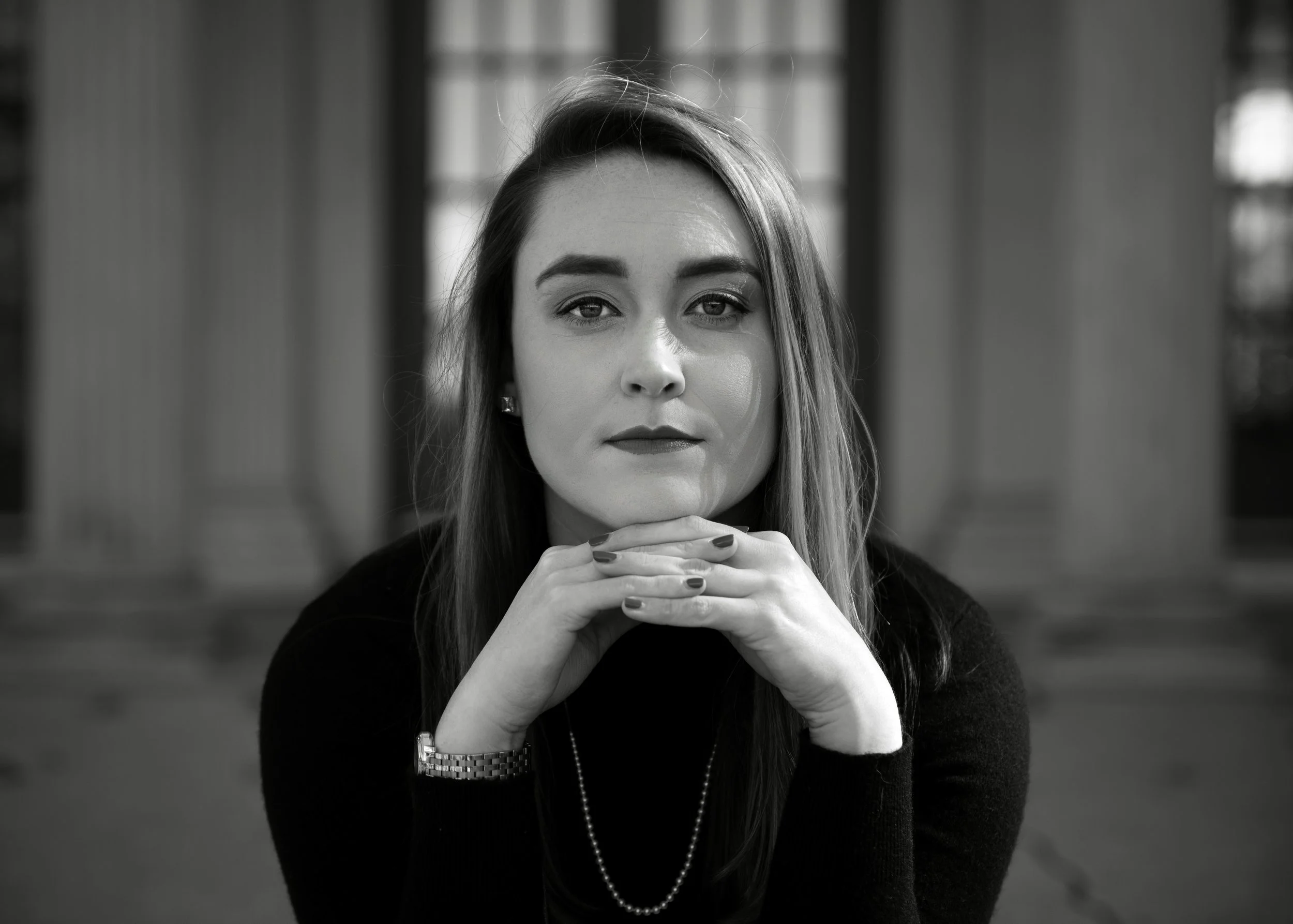 Black and white portrait of a professional woman with straight hair, wearing a black top and jewelry, sitting with her hands under her chin, looking directly at the camera in an outdoor setting.