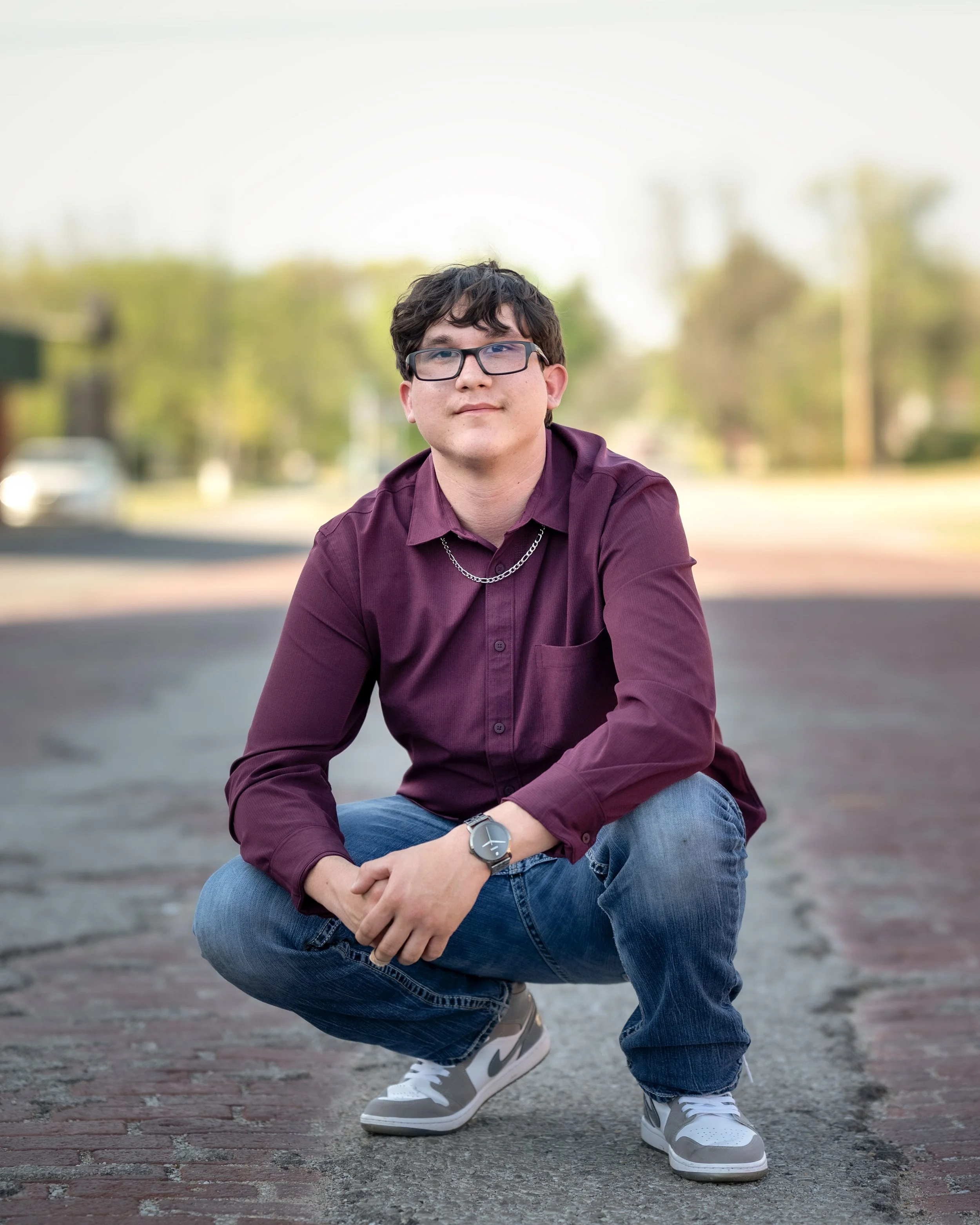 A young man with dark curly hair, glasses, wearing a maroon button-up shirt, blue jeans, and white sneakers, squatting on a brick-paved street with a blurred background of trees and cars.