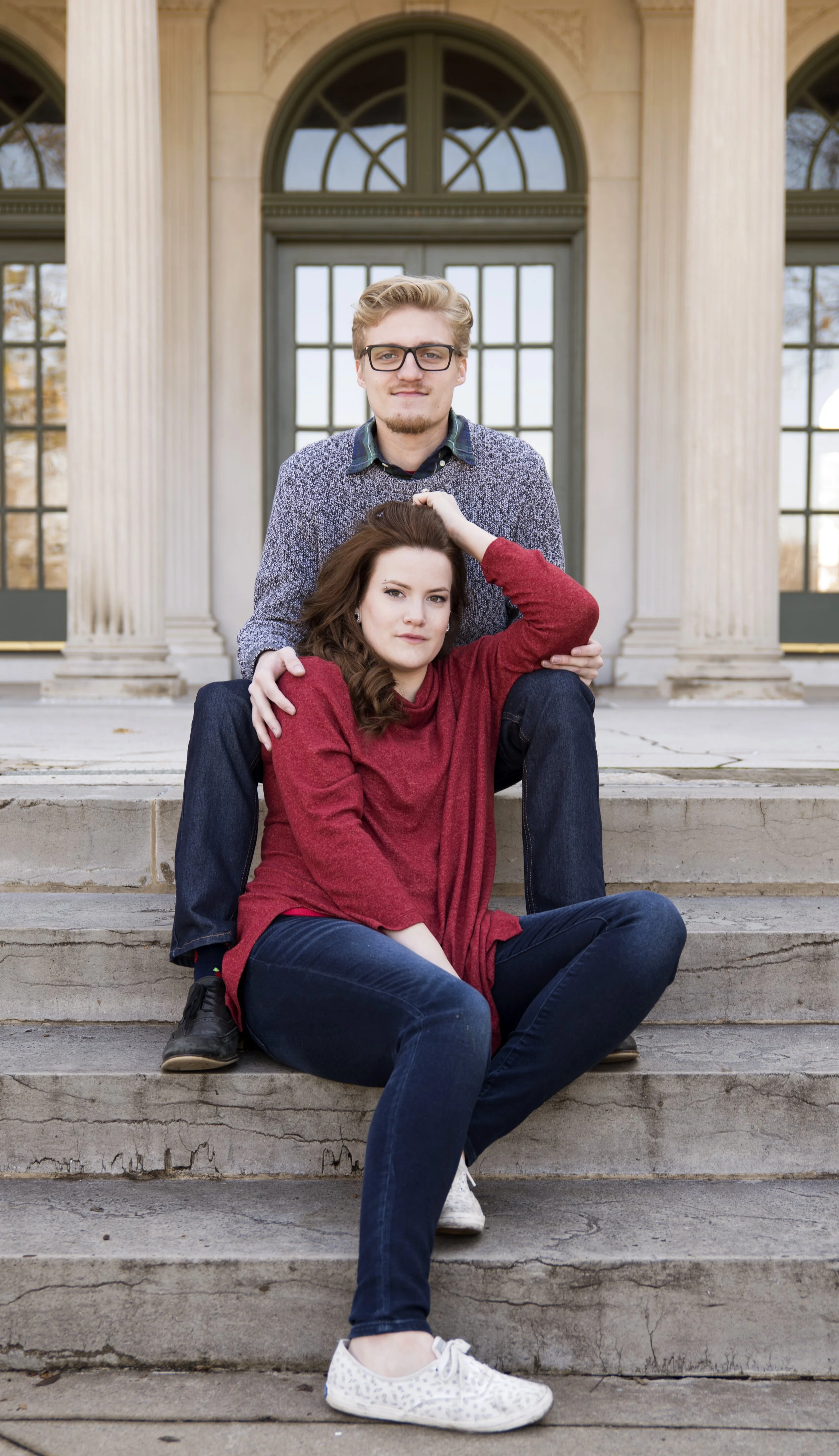 A young man and woman sitting on stone steps in front of a historic building with large columns and arched windows. The man is standing behind the woman, who is sitting with her legs crossed, and they are posing for a photo.