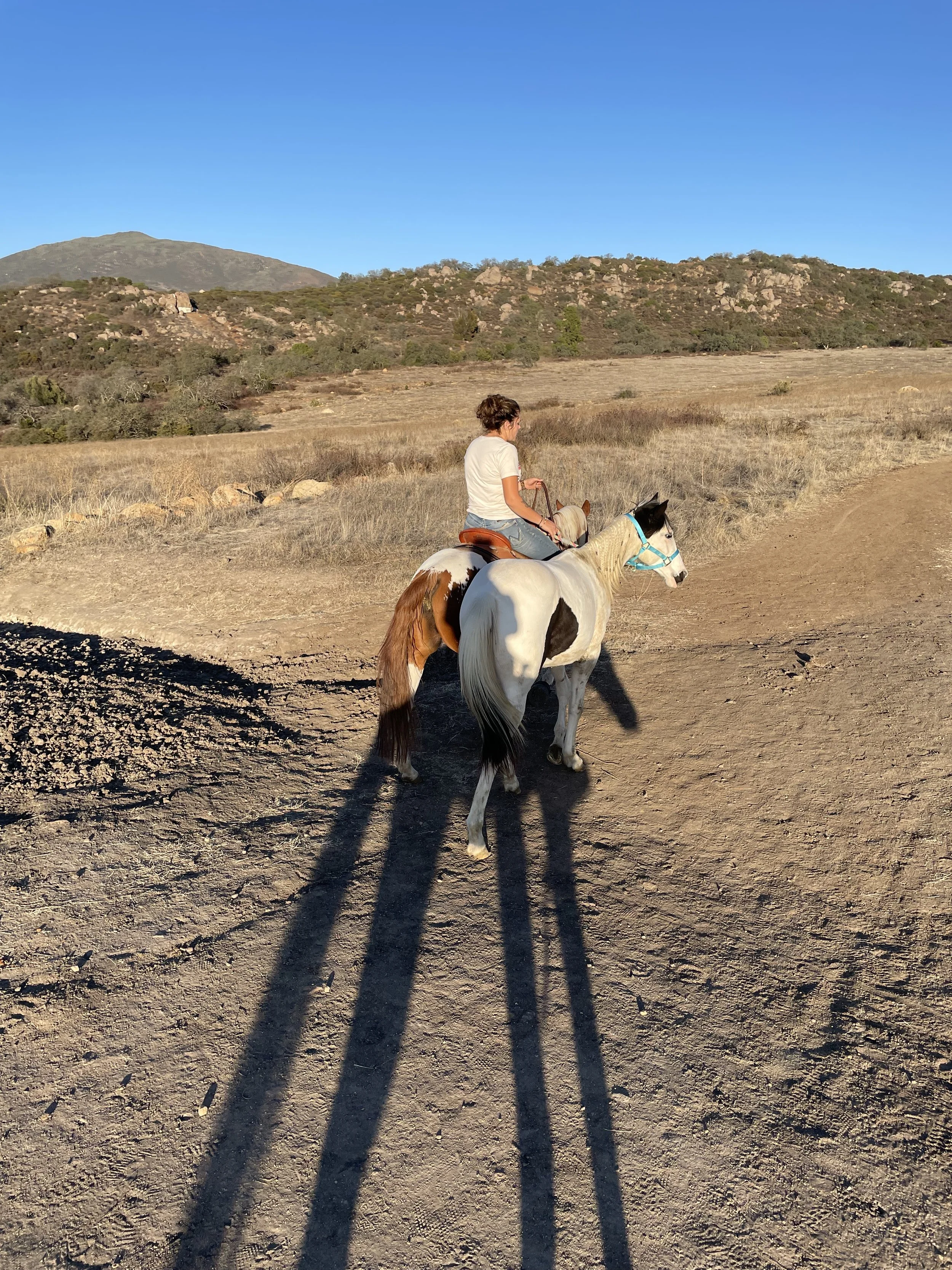 A woman riding a white and brown paint horse on a dirt trail in a dry, grassy landscape with hills in the background under a clear blue sky.
