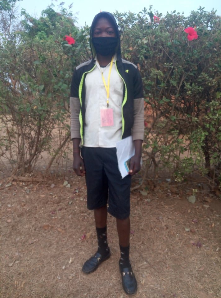 A young person standing outdoors in front of a bush with pink flowers, wearing a black mask, hoodie, white shirt, shorts, and sandals, holding papers and a smartphone.