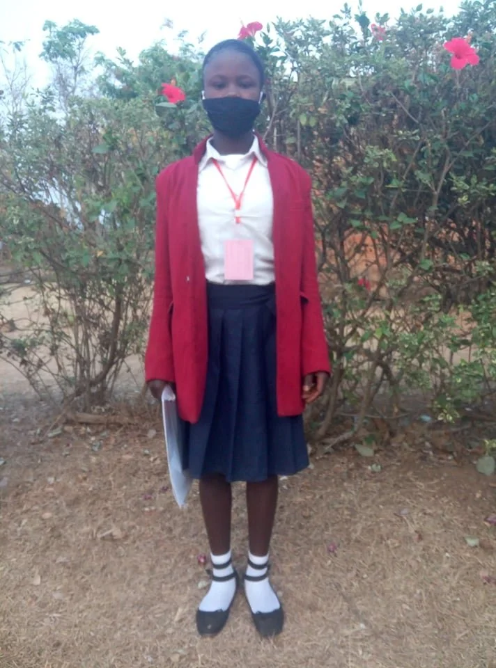 A young girl wearing a school uniform, red cardigan, white shirt, navy blue skirt, and black shoes with white striped socks stands outdoors in front of bushes with pink flowers. She is wearing a black face mask and holding a notebook or folder.