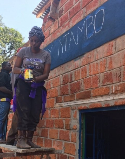A woman holding a jar, standing on a wooden platform outside a brick building, with a sign that reads 'Y NTAMBO' in the background.