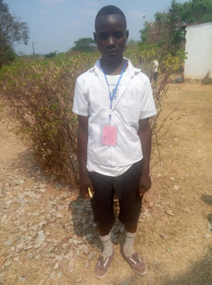 A young boy standing outdoors on a dirt ground with shrubbery in the background. He is wearing a white shirt, black shorts, gray socks, and brown slide sandals. He has a blue lanyard with a pink identification card around his neck and is holding a ye