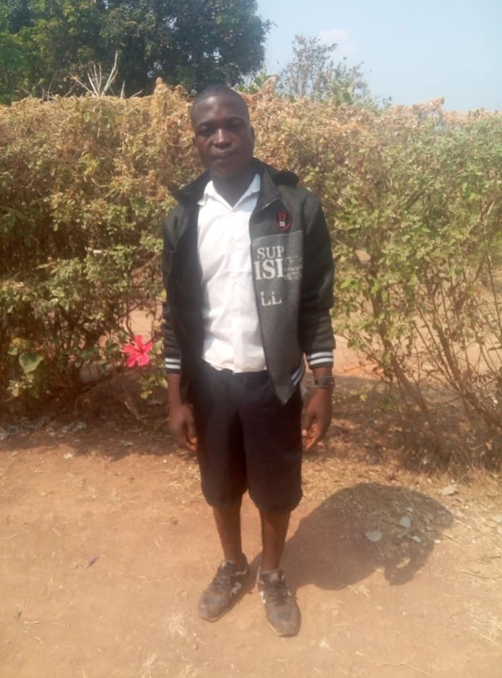 A young boy standing outdoors on dirt ground, wearing a white shirt, black shorts, a dark jacket, and sneakers, with bushes and a cloudy sky in the background.
