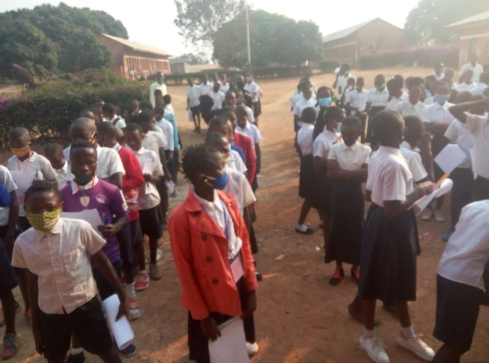 Students standing in lines outdoors at school, wearing masks and holding papers.