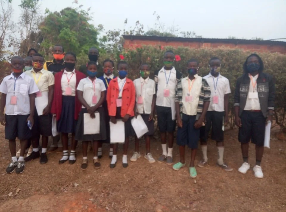 Group of students standing outdoors, wearing masks, some holding papers, with trees and a building in the background.