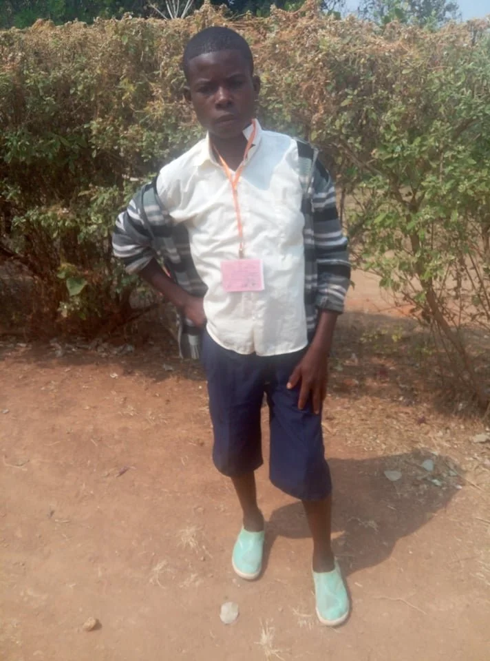 A young boy standing outdoors on dirt ground with bushes in the background, wearing a white shirt, navy shorts, and light-colored slip-on shoes, with a pink identification badge around his neck, and one hand on his hip.