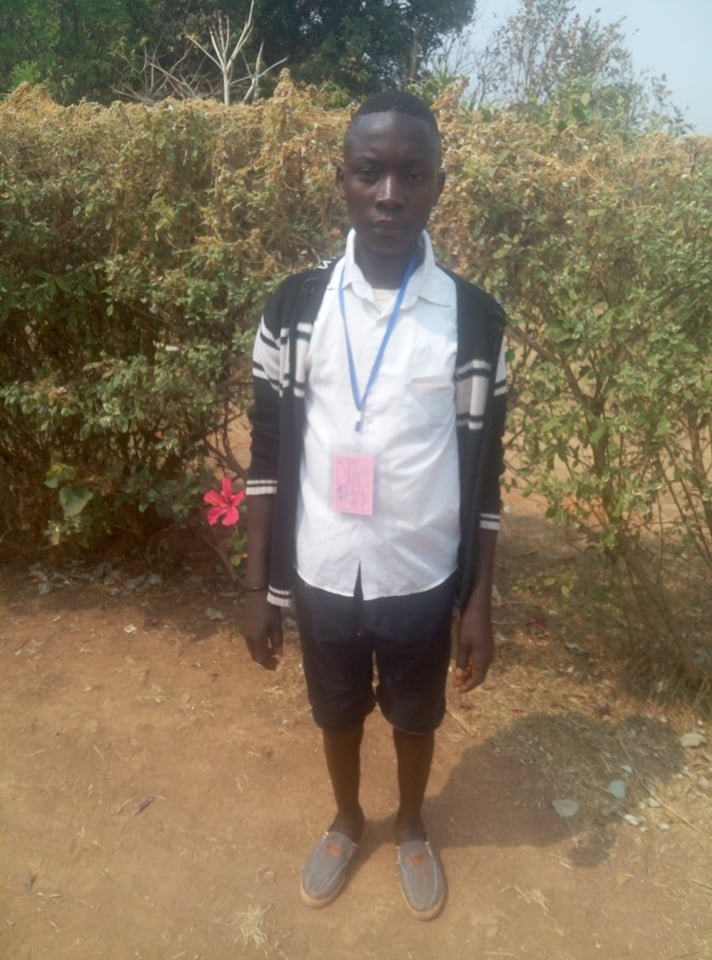 Young boy standing outdoors in front of bushes, wearing a white shirt, black shorts, a black and white striped cardigan, gray slip-on shoes, and a blue lanyard with an ID card.