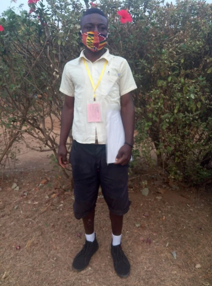 A young boy standing outdoors during daytime, wearing a patterned face mask, a light-colored short-sleeve shirt, dark shorts, white socks, and black shoes. He has a yellow lanyard with an identification badge and is holding papers in his left hand, w