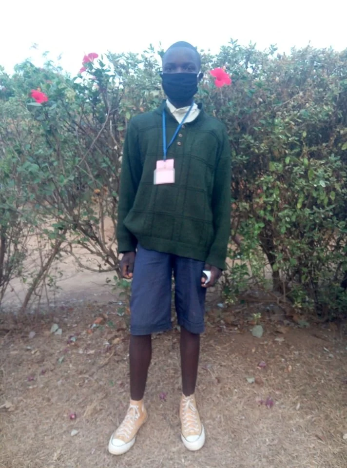A young man standing outdoors in front of bushes with pink flowers, wearing a black face mask, dark green jacket, gray shorts, high-top beige sneakers, and a lanyard with an ID card.