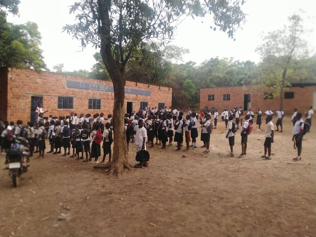 School students in uniform standing and waiting outside school buildings under trees on a dirt ground.