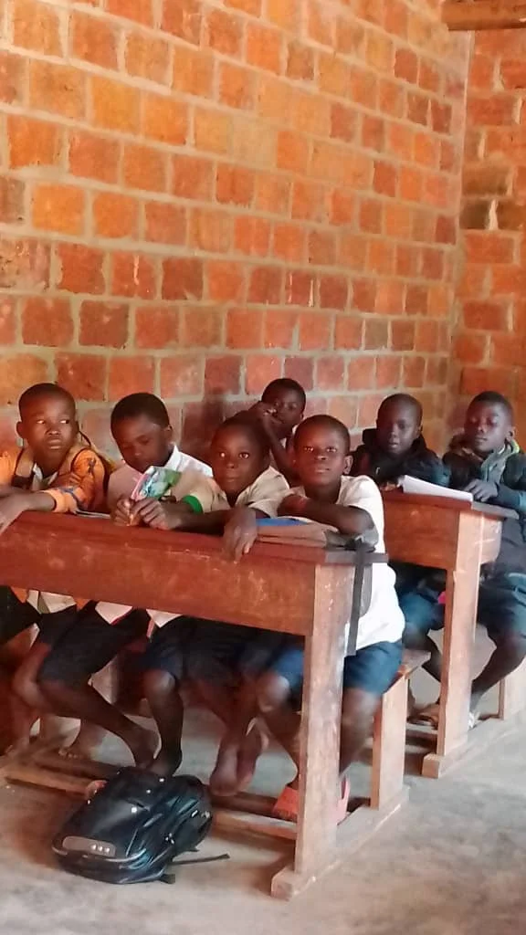 Young students sitting at wooden desks in a classroom with brick walls, looking toward the camera.
