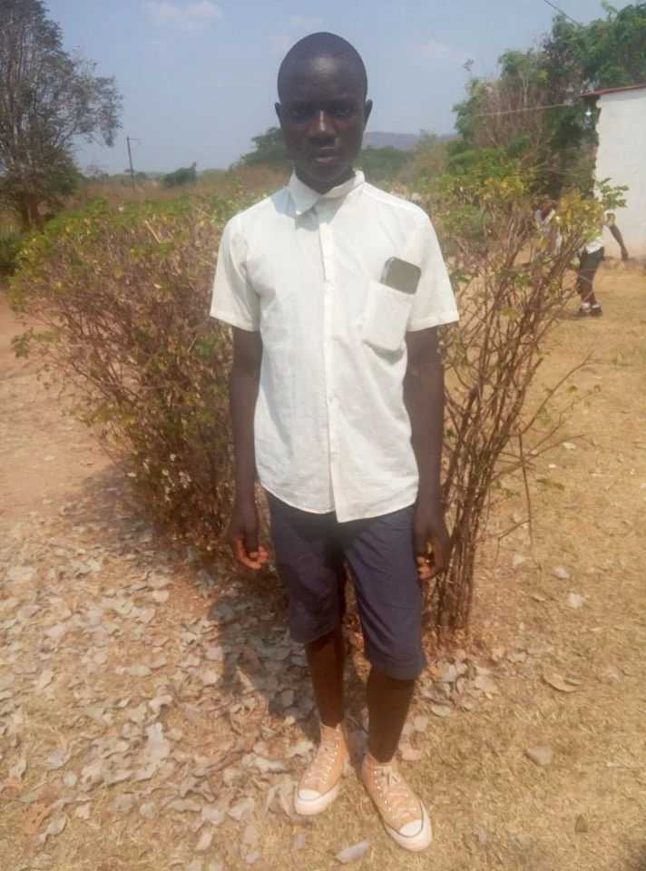 A young boy standing outdoors in front of leafless bushes, wearing a white short-sleeve shirt, dark shorts, and tan sneakers.