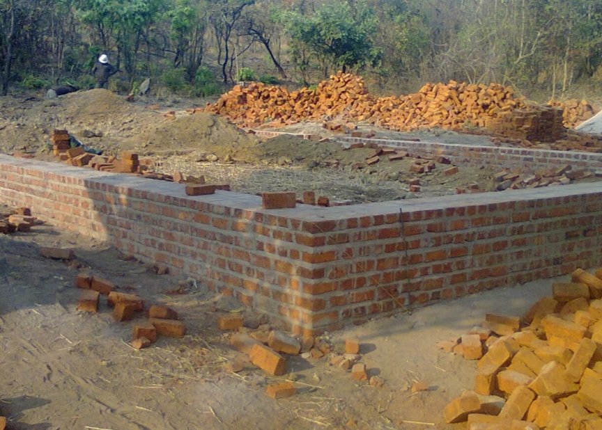 Construction site with a brick foundation and pile of bricks in the background, surrounded by dirt and trees.